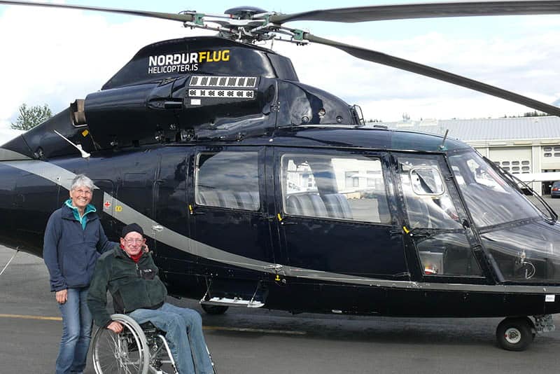 Een echtpaar waarvan 1 persoon in een rolstoel poseren bij de helicopter voor hun rondvlucht