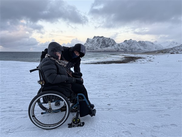 Persoon in rolstoel met iemand ernaast in de sneeuw met bergen en zee op de achtergrond