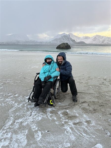 een vrouw in een rolstoel met een zittende man ernaast op een bevroren strand en bergen op de achtergrond