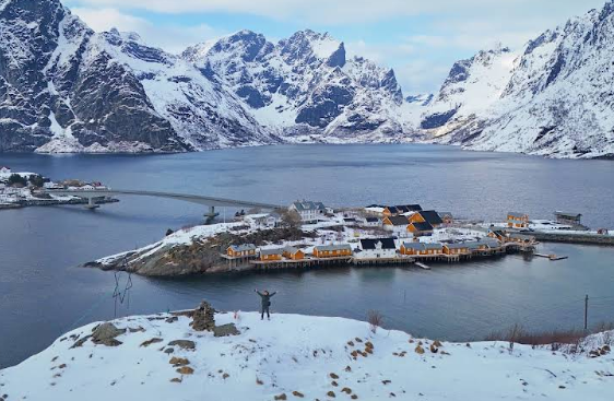 Man zwaait naar de camera in een sneeuwlandschap met water en bergen en en dorpje