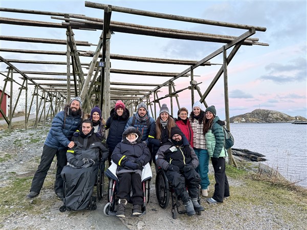 Groep mensen, sommigen in een rolstoel, poseren bij visdroogrekken op de Lofoten