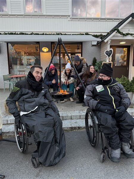2 mensen in een rolstoel op straat met erachter mensen die een vuurschaal aansteken voor een winkel