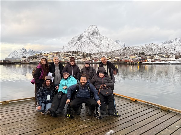 Groep mensen waarvan 2 in een rolstoel op een steiger bij een stad aan het water met besneeuwde bergen op de achtergrond