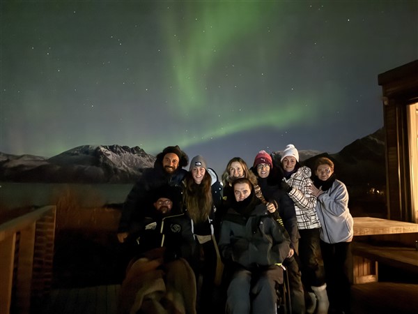 Groep mensen, sommige in rolstoel, buietn bij een huis met erboven noorderlicht en erachter bergen