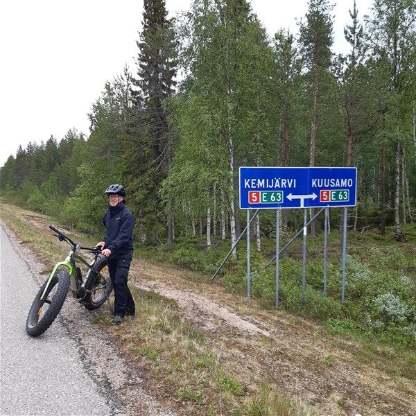 Een man met een fatbike aan de kant van de weg met op de achtergrond een bos en een verkeersbord