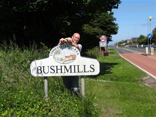 Man kijkt lachend vanachter een plaatsnaambord in Ierland