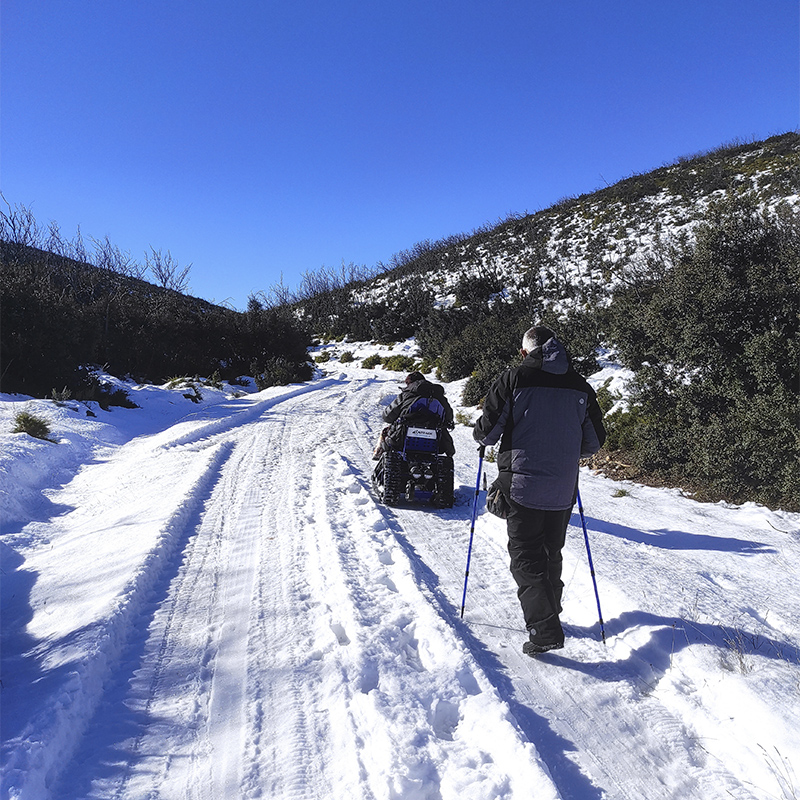 Een rolstoelgebruiker in een aangepaste rolstoel rijdt over een besneeuwd bergpad samen met een wandelaar