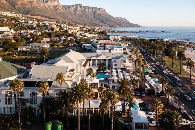 Een groot wit hotel met een zwembad en parasols langs een weg en strand aan zee met bergen inde achtergrond en bebouwing op de heuvelflank